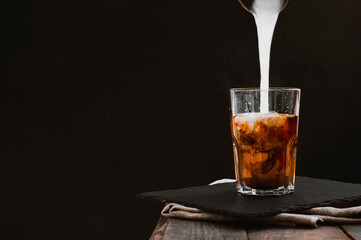 Pouring milk in cup of coffee on a wooden table in black background. Glass cup Iced coffee