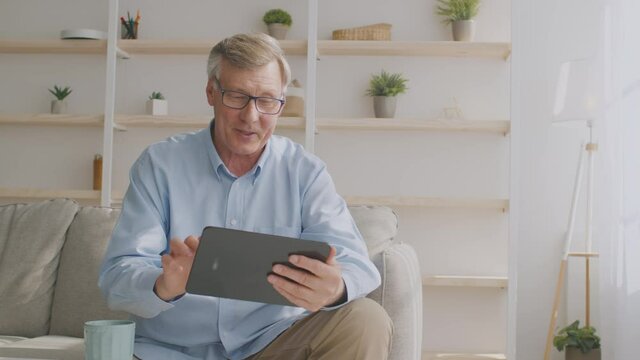 Cheerful senior man using digital tablet online, enjoying internet and gadget at home, tracking shot, empty space