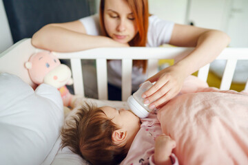 Obraz na płótnie Canvas Front view of caucasian woman mother taking care of his newborn baby child two months old bottle feeding with breast or formula milk while child is lying in cradle at home selective focus copy space