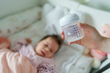Close up selective focus on empty baby bottle in hands of unknown woman mother holding it above the bed at home with sleeping infant in background - newborn feeding nursing concept concept copy space