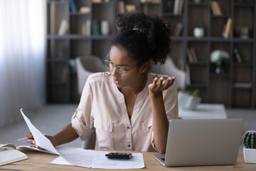 Stressed millennial generation african mixed race woman in eyewear looking at financial documents...