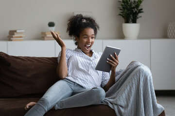 Emotional happy young african ethnicity multiracial woman reading unexpected good news on touchpad, feeling overjoyed at home. Excited millennial generation mixed race lady celebrating online victory.
