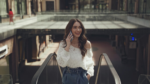cheerful young woman talking on smartphone while standing with hand in pocket near escalator.
