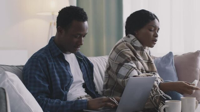 Young black man working on laptop at home, his sick wife sneezing and blowing nose, sitting wrapped in plaid on sofa