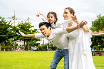 Portrait of asian happy family of three having fun together in park, little daughter sitting on father back and mother smile walking and having fun moments good time in summer park at home