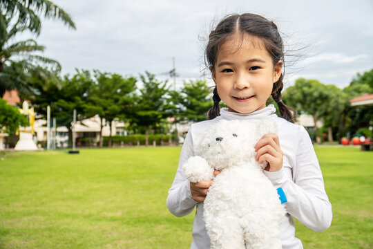 Happy asian little child girl standing in the grass and playing with her favorite teddy bear toy outdoors in summer park