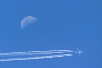 Fototapeta premium An aircraft and the a half moon in a blue sky. The aircraft is leaving behind lines of vapour trails. (Contrails) 