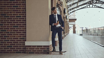 full length of businessman in glasses and suit holding coffee to go while posing near penny board in shopping mall.