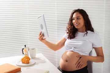 Happy pregnant young beautiful woman talking to mom using video call during morning breakfast. Concept of communication and positive attitude during pregnancy