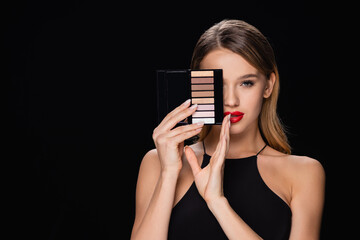 young woman obscuring face with eye shadows palette isolated on black.