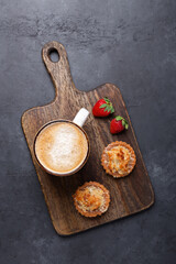 Cup of coffee, cakes and strawberries on a wooden cutting board on dark stone background. Top view