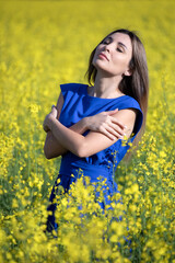Naklejka na ściany i meble Caucasian girl in a blue dress enjoys nature on a yellow rapeseed field. Countryside.
