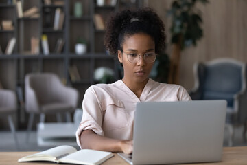 Concentrated young african woman in eyeglasses working on project on computer, using corporate software application in modern office. Focused smart biracial female student studying distantly at home.