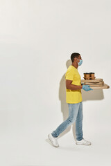 Full length shot of delivery man wearing protective mask and gloves looking away, carrying pizza boxes and drinks isolated over gray background