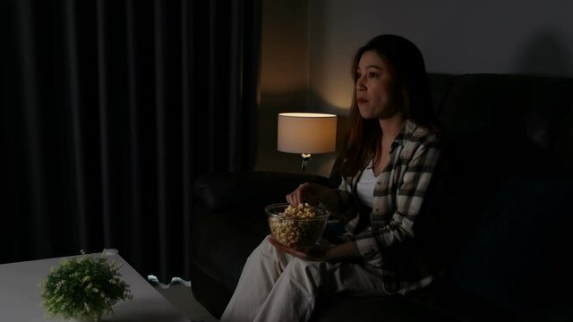 Funny young woman watching TV and eating popcorn on sofa at night