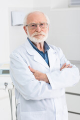 senior dentist in white coat standing with crossed arms in dental clinic.