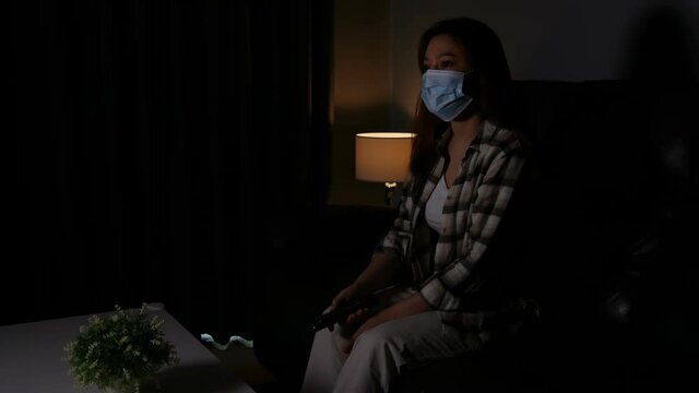 Young woman watching TV on sofa and wearing medical mask to protect coronavirus (Covid-19)