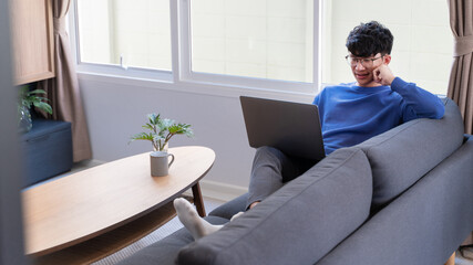 young attractive asian man relaxing comfortably working on laptop in bright living room at his home.