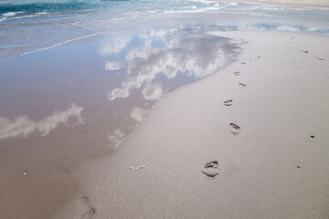 beach wet sand with fresh human bare footprints and blue cloud sky reflection and water waves