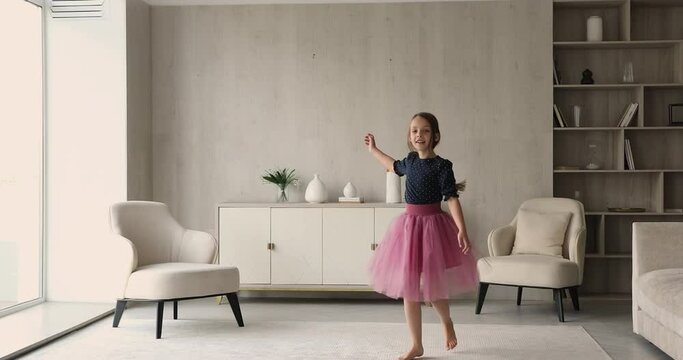 Little beautiful girl wear pink fluffy skirt dancing barefoot on carpet in modern cozy living room. Pretty kid listens music jumping having fun alone at home. Hobby, dance school for children concept