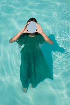 woman in green dress in swimming pool holding round mirror hiding her face