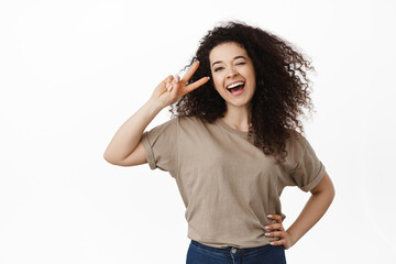 Portrait of happy and positive brunette woman, laughing and smiling, showing peace v-sign near eye, kawaii pose, standing against white background