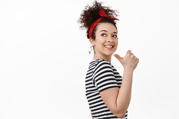 Portrait of young woman, female rights activist, pointing finger left behind her shoulder, smiling and looking hopeful, standing against white background