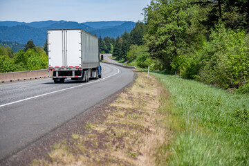 Blue big rig semi truck tractor transporting cargo in dry van semi trailer running for delivery on the winding highway road in a picturesque region with green forest and mountains