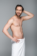 happy and muscular man wrapped in white towel standing with hand on hip isolated on grey.