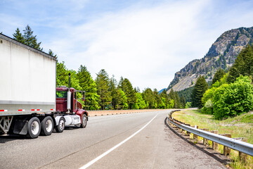 Burgundy day cab big rig semi truck transporting cargo in dry van semi trailer running on the turning mountain highway road