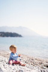 Small child sits on a pebble beach near the water, holds a stick in his hand and looks at the sea