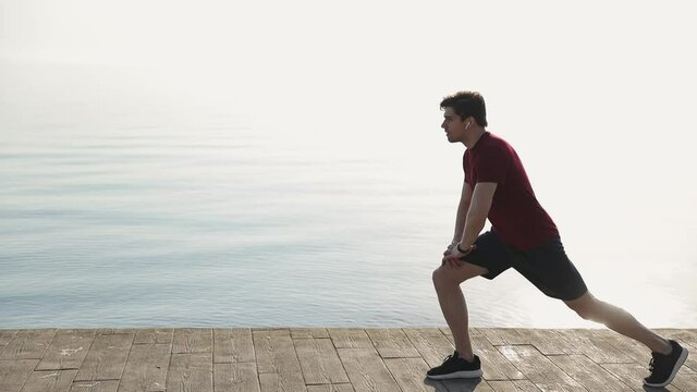 Young strong sporty athletic sportsman man 20s wear red t-shirt black shorts warm up training doing stretch exercise for legs at sunrise over sea beach ocean outdoor jog on pier seaside in summer day
