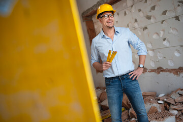 Architect with yellow safety helmet, blue shirt and jeans checks construction progress on building site in loft, attic with a yellow  folding rule