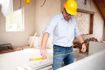 Architect with yellow safety helmet, blue shirt and jeans checks construction progress on building site in loft, attic with a yellow  folding rule