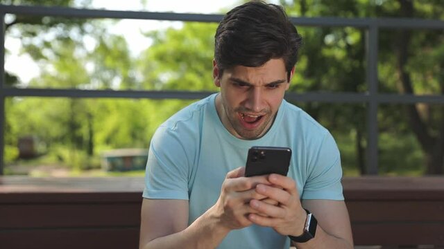 Young calm man 20s wears blue t-shirt sit on bench hold in hand using mobile cell phone read sms rest relax in spring green city park sunshine day outdoors on nature. Urban lifestyle leisure concept