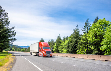 Energy saving design big rig red semi truck transporting cargo in dry van semi trailer moving on the divided highway road with green summer trees