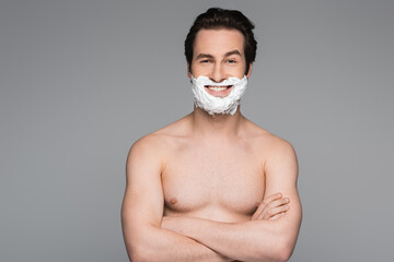 happy and shirtless man with shaving foam of face posing with crossed arms isolated on grey.