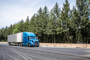 Stylish blue big rig long haul industrial semi truck transporting cargo in dry van semi trailer running on the highway road with pine trees on the side © vit