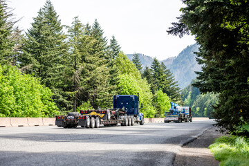 Team of two blue big rig semi trucks with step down semi trailers transporting transport equipment running at the convoy on the green forest road