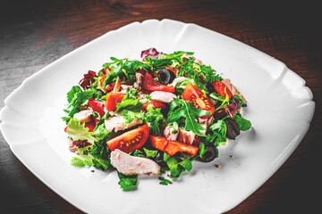Fresh salad with chicken breast, arugula, black olives,red pepper, lettuce, fresh sald leaves and tomato on a white plate on wooden table background