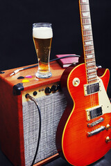 Amplifier for guitar with guitar, glass of beer and notepad on black background.