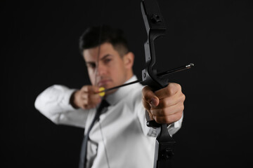 Businessman with bow and arrow practicing archery against black background, focus on hand