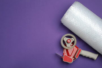 Bubble wrap roll and tape dispenser on purple background, flat lay. Space for text