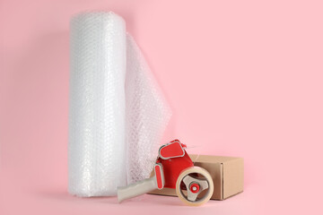 Bubble wrap roll, tape dispenser and cardboard box on pink background, space for text
