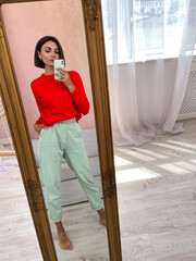 Stylish women at home in bright red sweater and mint green pants take photo selfie in mirror on phone for stories social media, vertical