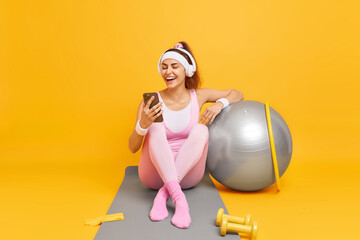 Happy young fitness woman ready to start training looks gladfully at smatphone display laughs positively wears wireless headphones listens audio track while having workout takes break in gym - obrazy, fototapety, plakaty