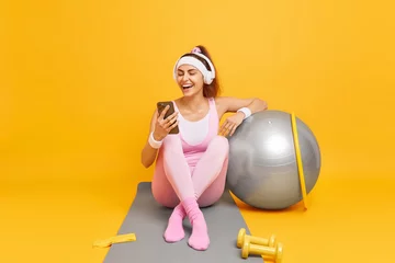Foto op Canvas Happy young fitness woman ready to start training looks gladfully at smatphone display laughs positively wears wireless headphones listens audio track while having workout takes break in gym © Wayhome Studio