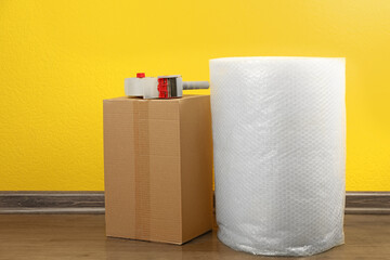 Bubble wrap roll, tape dispenser and cardboard box on floor near yellow wall