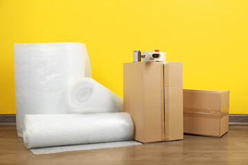 Bubble wrap rolls, tape dispenser and cardboard boxes on floor near yellow wall - obrazy, fototapety, plakaty