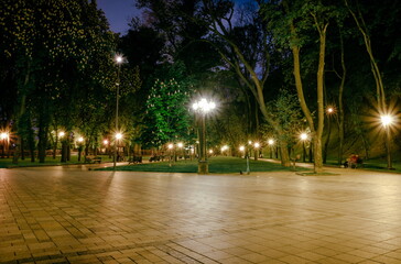 Obraz premium The tiled road in the night green park with lanterns in spring. A benches in the park during the spring season at night. Illumination of a park road with lanterns at night. Mariinsky Park. Ukraine